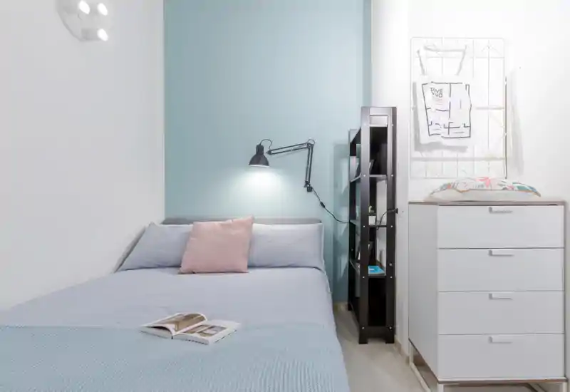 Camera da letto luminosa e minimal con letto ben rifatto davanti a una parete azzurra, lampada da lettura, scaffalatura e comò bianco — ambiente pulito e accogliente ideale per un soggiorno riposante.