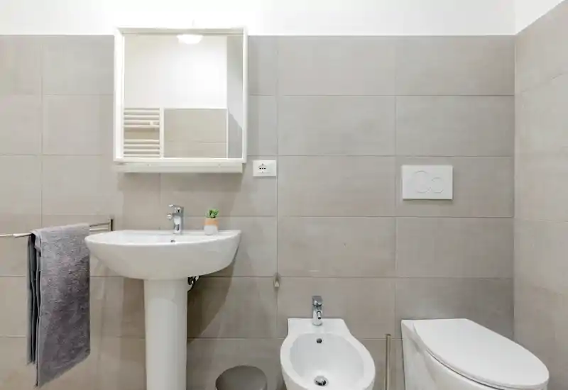 Clean, modern bathroom with a pedestal sink, wall-mounted mirror cabinet, bidet and toilet in neutral grey tiles. Bright, minimal and well-lit space suitable for guest or ensuite use.