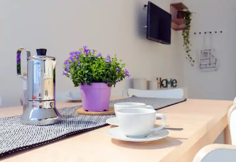 Bright, styled dining table with cups, a moka pot and a potted plant on a runner — ideal for showcasing a cozy breakfast setup.