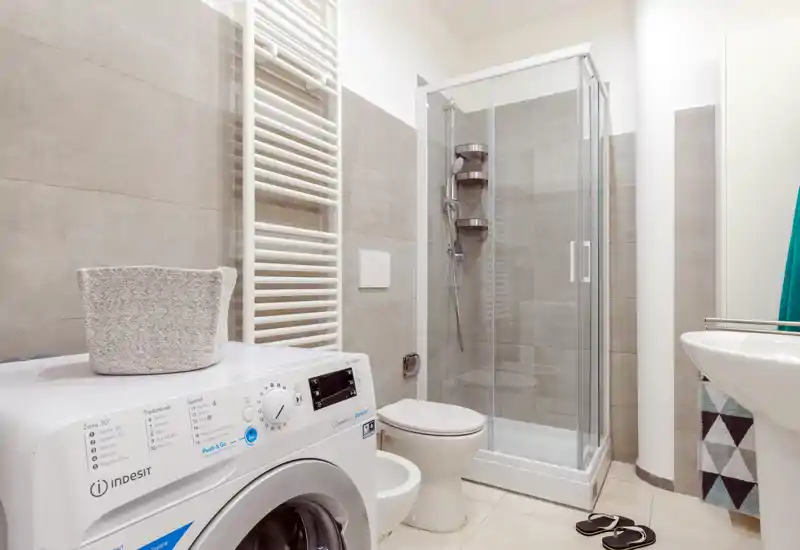 Clean modern bathroom with a corner glass shower, toilet and a front-loading washing machine; neutral tiles and bright lighting create a fresh, functional space.