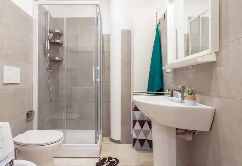 Bright, modern bathroom with a glass shower enclosure, wall-mounted toilet, and a pedestal sink with mirror; neutral tiles and clean design.