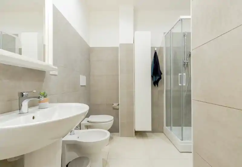 Modern, compact bathroom with a pedestal sink, bidet, toilet and glass shower cubicle; neutral beige tiles and clean lines create a fresh, minimal look.