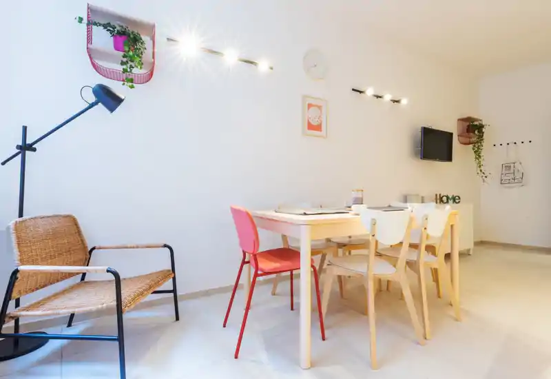 Bright, modern living/dining area with a light wood table, mixed chairs (including a red accent chair), wicker armchair, wall-mounted TV and decorative plants — a stylish, airy space ideal for relaxed meals or socialising.
