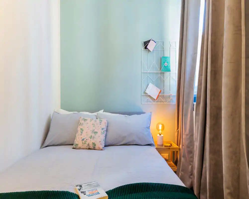 Dormitorio luminoso y acogedor con cama doble, cojín decorativo y lámpara de mesita cálida junto a una ventana con cortinas.