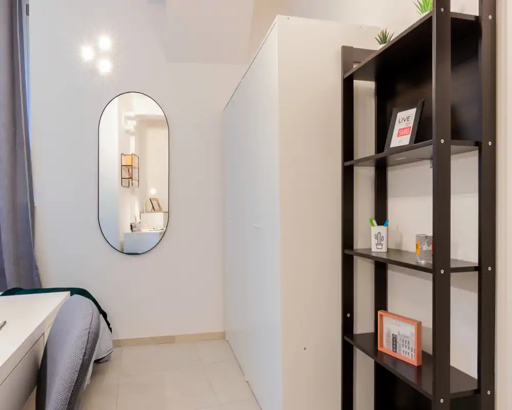 Rincón de dormitorio luminoso y compacto con el borde de la cama visible, armario y estantería; decoración minimalista y buena luz.