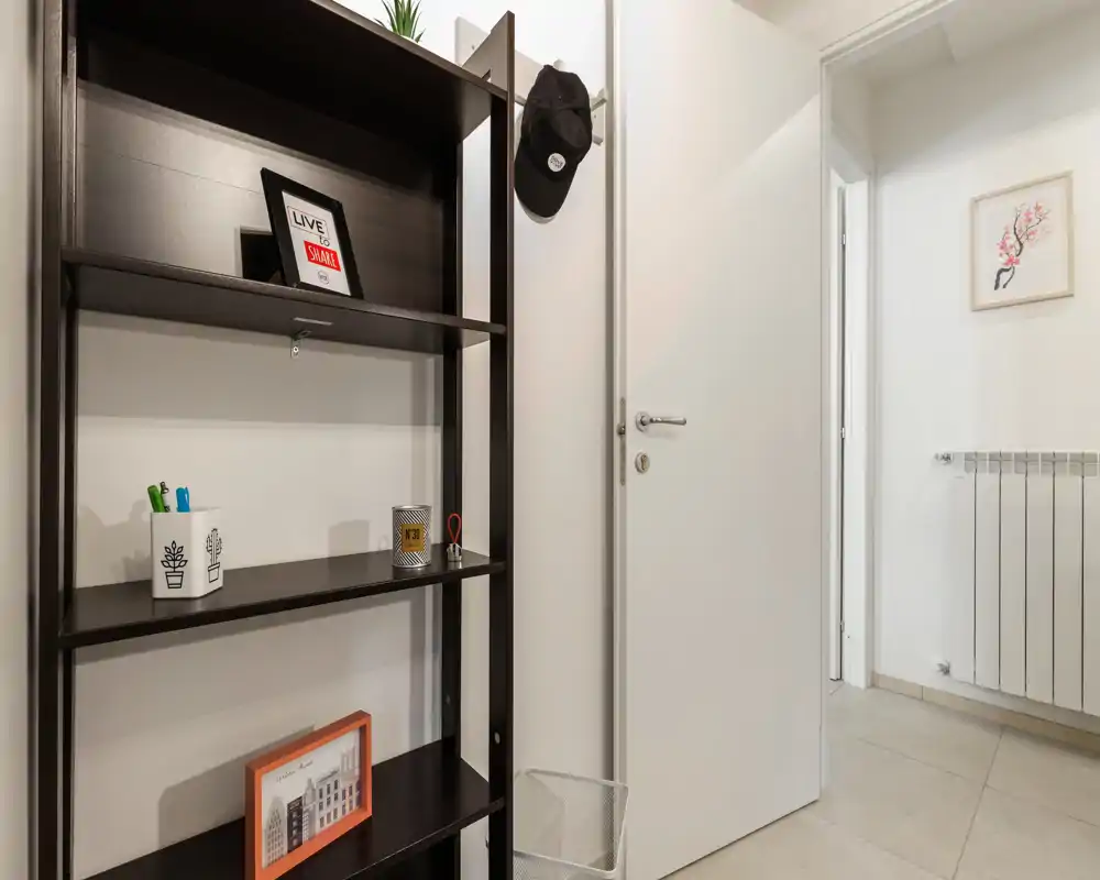 Entrada compacta y bien iluminada con una estantería estrecha, perchas para gorra y una puerta blanca que da a un pasillo luminoso: recibidor minimalista y ordenado, ideal para alquiler urbano.