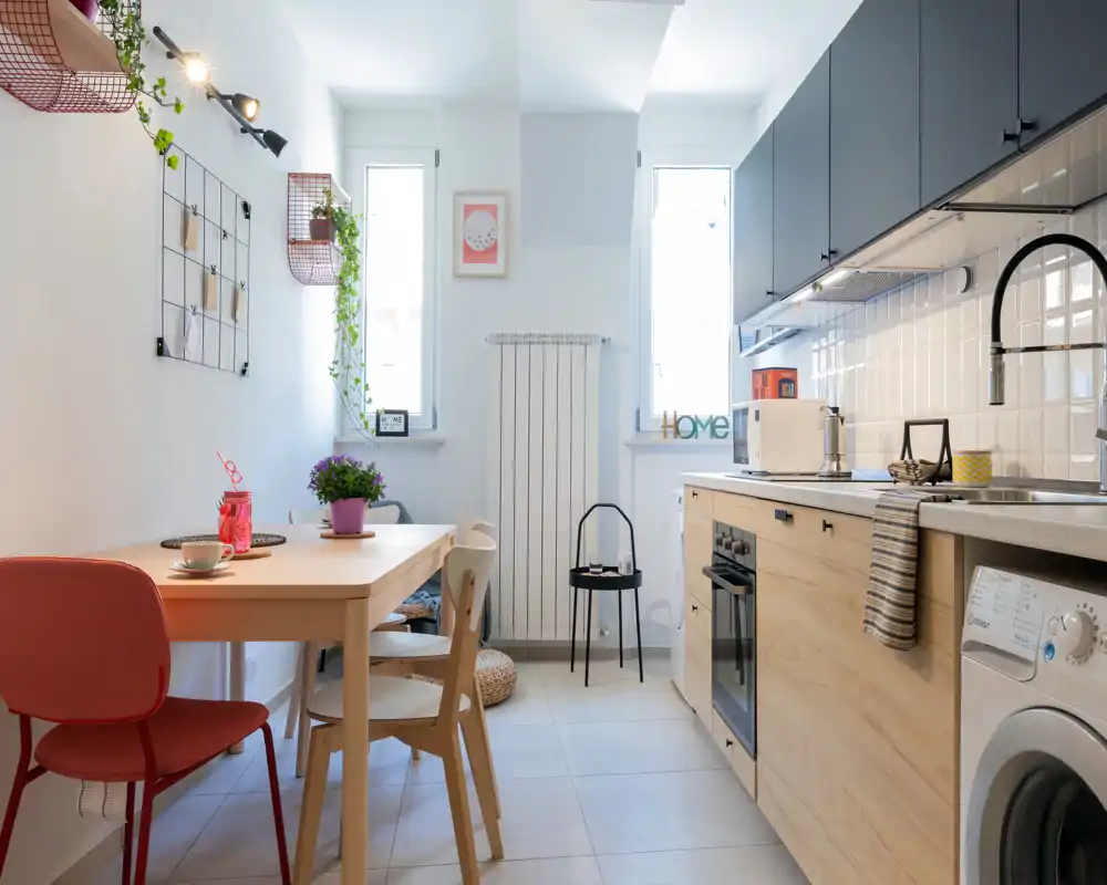 Cocina moderna y luminosa con muebles bajos de madera, armarios superiores oscuros, horno integrado y lavadora. Una pequeña mesa de comedor con sillas mixtas, plantas y detalles decorativos crean un espacio acogedor y funcional.