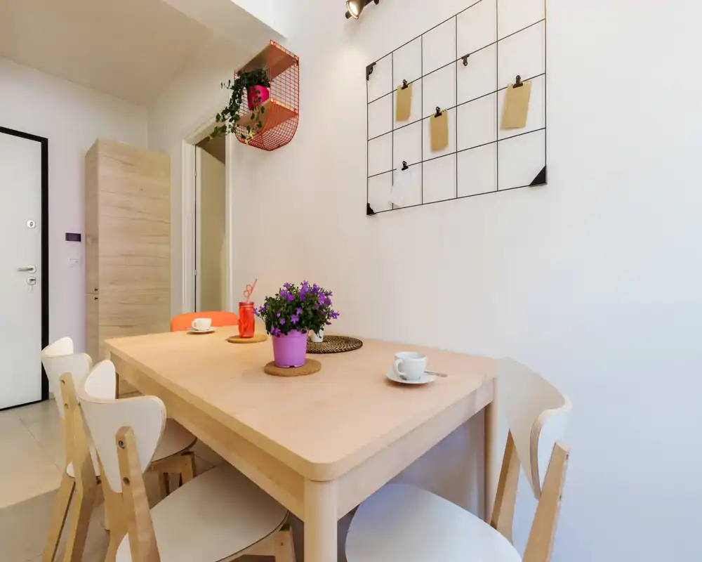 Zona de comedor luminosa y moderna con mesa de madera clara, sillas sencillas y una pequeña planta en maceta que crea un ambiente acogedor y minimalista.