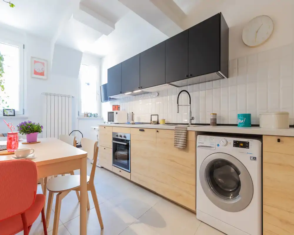 Cocina moderna y luminosa con muebles bajos de madera clara, encimeras con armarios superiores negros, horno integrado y lavadora frontal; una mesa de madera con sillas coloridas y plantas aporta un ambiente acogedor y bien iluminado.