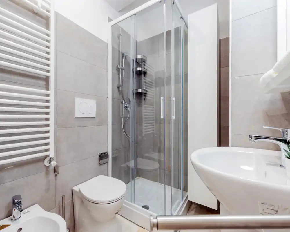 Baño moderno y compacto con ducha acristalada, inodoro suspendido, bidé y lavabo; azulejos neutros y un armario alto que aportan una imagen limpia y luminosa.