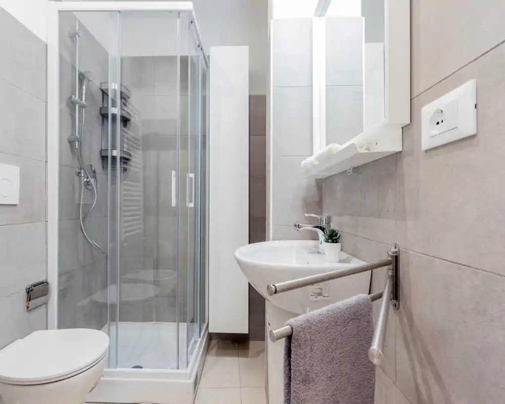 Baño moderno y bien iluminado con mampara de ducha de vidrio, lavabo mural, inodoro y acabados en azulejos neutros — limpio y funcional.