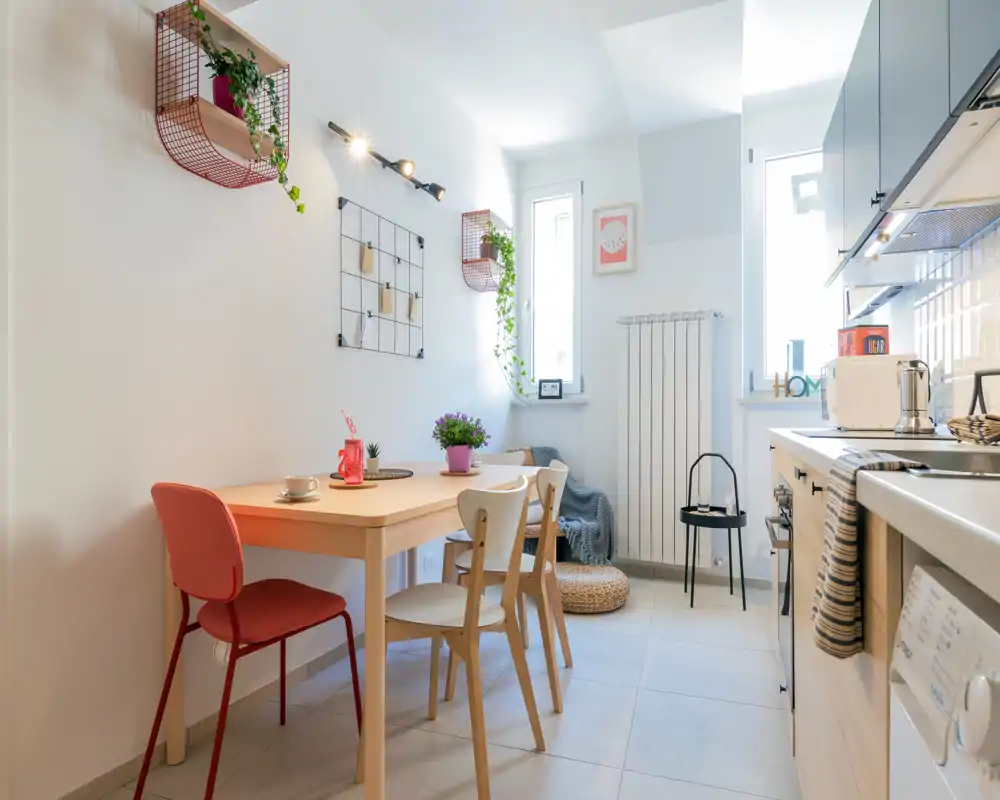 Atractiva zona de comedor integrada en cocina compacta: mesa de madera con sillas mezcla, plantas y detalles decorativos que crean un espacio luminoso y acogedor para comidas informales.