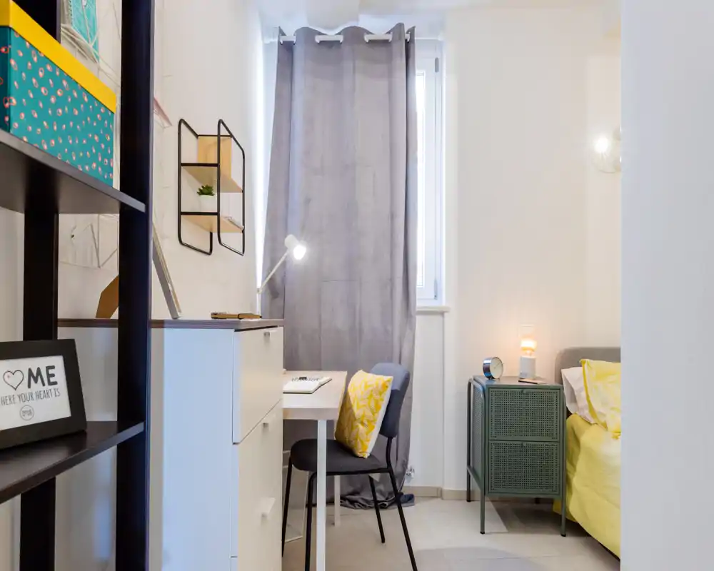 Rincón de dormitorio luminoso y compacto con cama de ropa de cama amarilla, mesita con lámpara y pequeño escritorio junto a la ventana — ideal para una habitación acogedora o de estudiante.