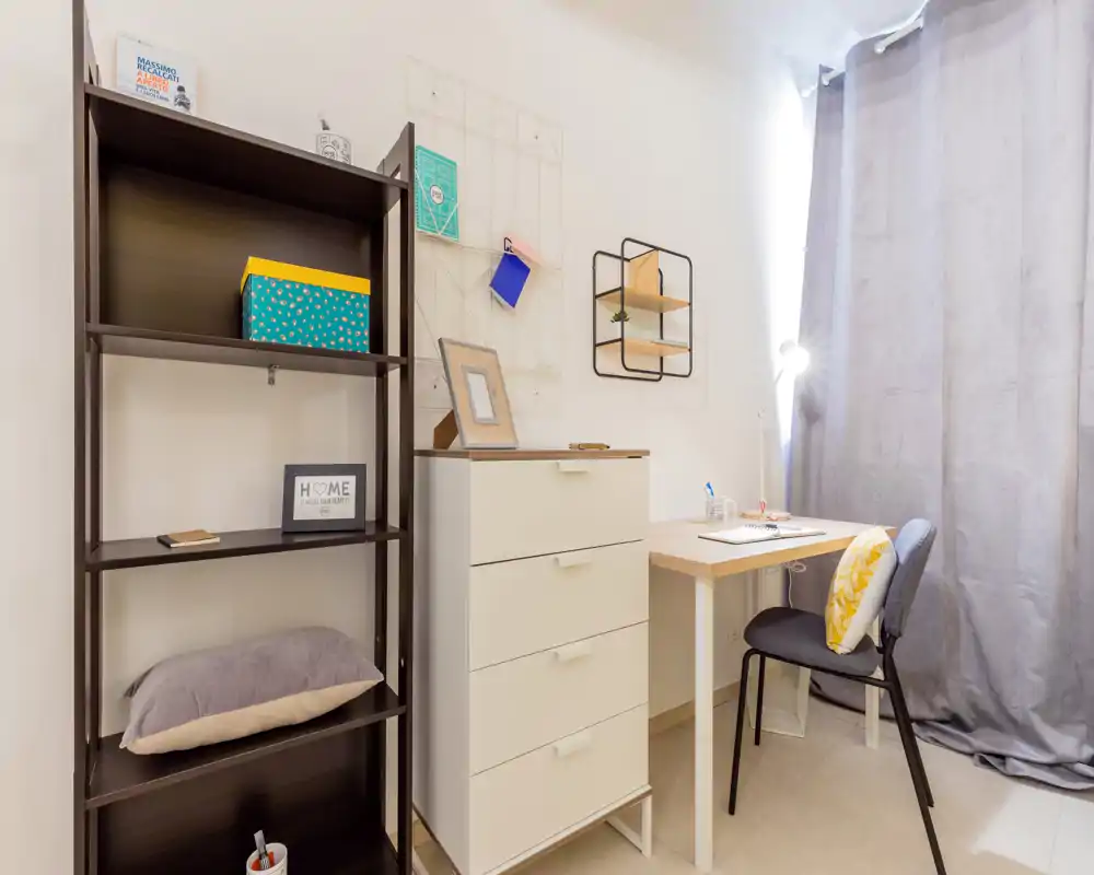 Rincón de dormitorio compacto con estantería, cómoda y escritorio junto a una ventana con cortina; luminoso, ordenado y decorado con detalles sencillos.