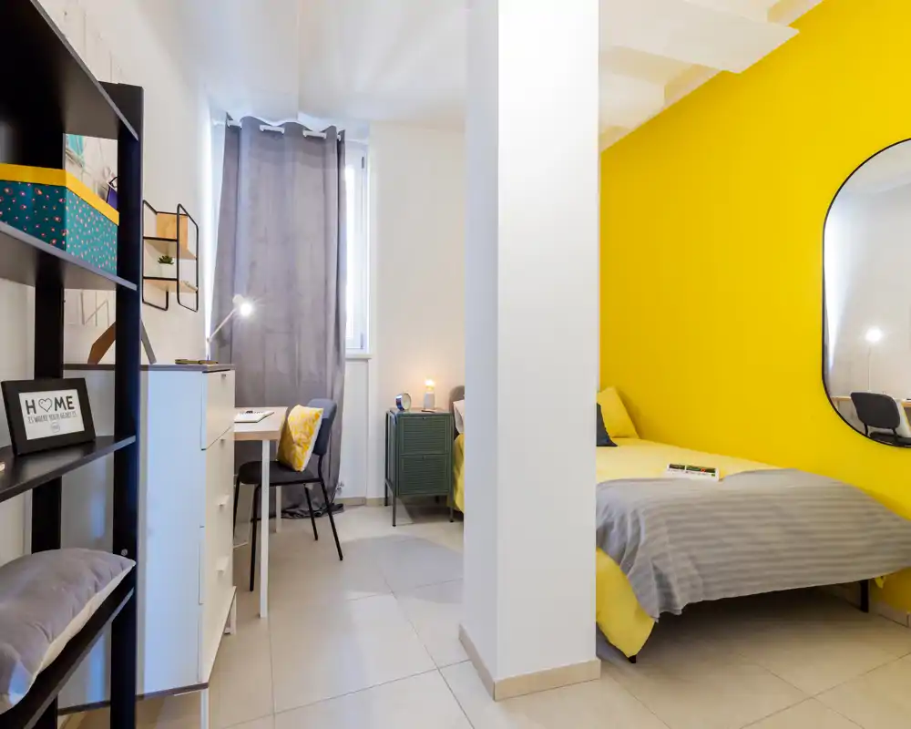 Dormitorio moderno y luminoso con pared amarilla intensa, cama cómoda, mesita con lámpara y un pequeño espacio de trabajo junto a la ventana.