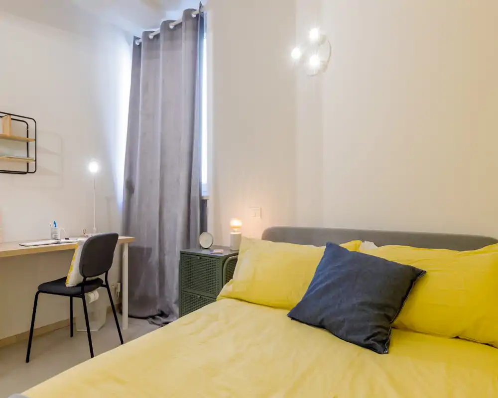 Dormitorio luminoso y bien decorado con una amplia cama con ropa de cama amarilla, mesita con lámpara y un pequeño escritorio junto a la ventana — perfecto para descansar y trabajar.