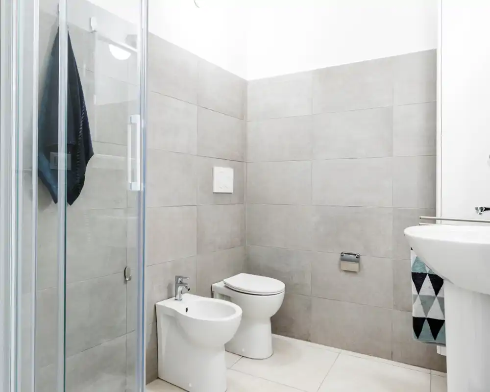 Baño moderno y luminoso con ducha, bidé e inodoro, azulejos grises neutras y lavabo de pedestal. Diseño limpio y minimalista con buena luz.