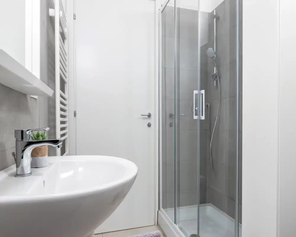 Baño contemporáneo y compacto con mampara de ducha de vidrio en esquina y lavabo de cerámica blanca; azulejos neutros y grifería limpia para un aspecto moderno y minimalista.