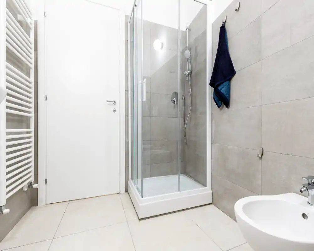 Baño moderno y minimalista con mampara de ducha de vidrio, bidé mural, radiador toallero y azulejos grandes de tonos neutros — brillante y limpio.