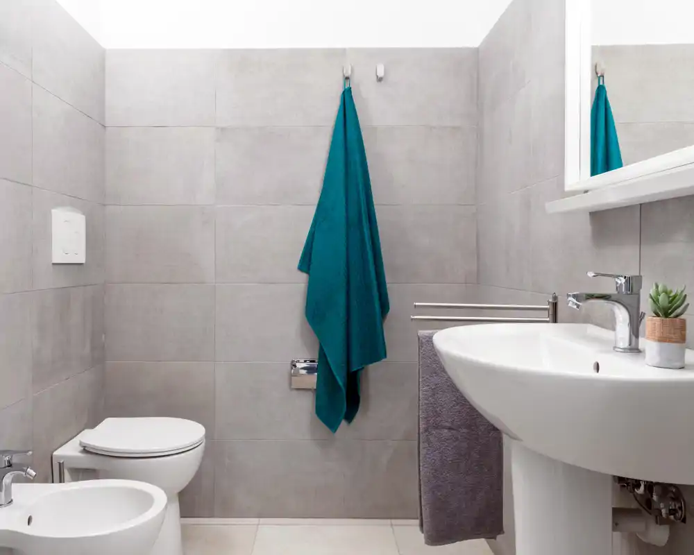 Baño moderno y luminoso con inodoro empotrado, bidé y lavabo blanco. Azulejos grises y una toalla turquesa crean un aspecto limpio y contemporáneo.