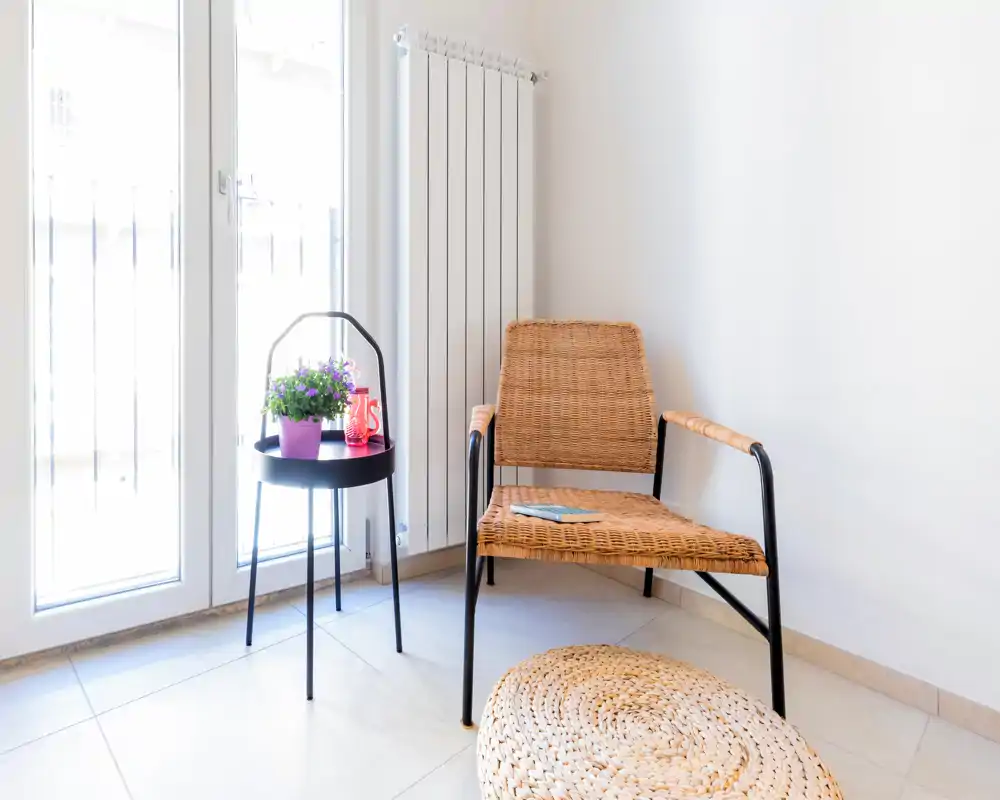 Rincón de asiento luminoso y minimalista con una silla de mimbre, una mesita y un puf trenzado junto a una gran puerta de cristal que aporta abundante luz natural.