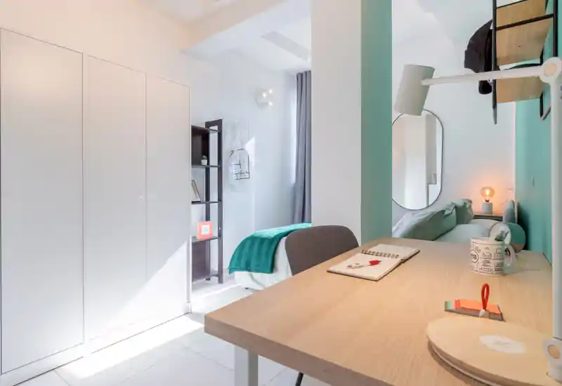 Dormitorio moderno y luminoso con cama bien hecha, escritorio en primer plano y estantería elegante. Luz natural, tonos pastel y una manta acogedora crean un ambiente ideal para alquileres de corta estancia.