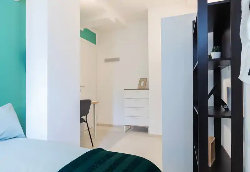 Rincón de dormitorio moderno y luminoso con cama bien hecha en primer plano, un pequeño escritorio y una cómoda al fondo, y estanterías contemporáneas a un lado.