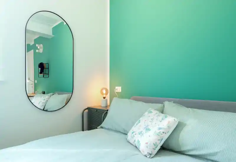 Dormitorio luminoso y con estilo con pared verde, cojines estampados y una lámpara de mesilla acogedora; la cama es el elemento principal y la habitación resulta fresca y atractiva.