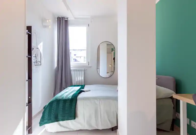 Dormitorio moderno y luminoso con cama doble centrada, ropa de cama clara y una manta verde; luz natural por la ventana y un espejo oval aumentan la sensación de espacio.