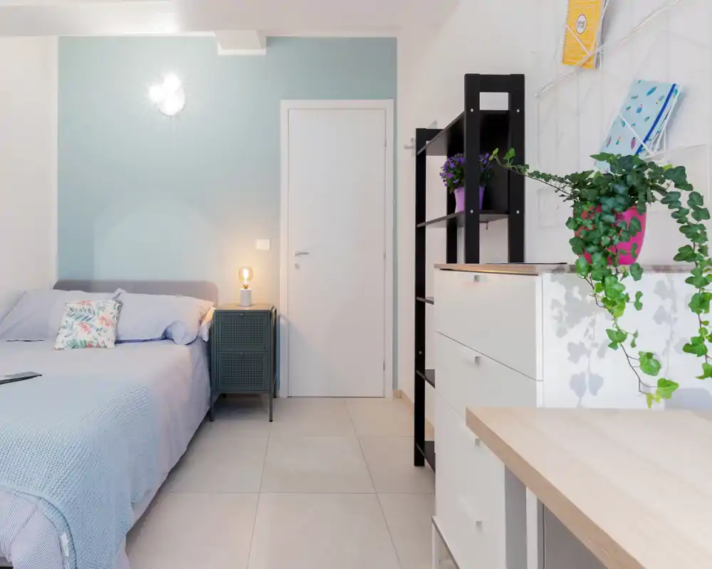 Dormitorio moderno y luminoso con cama bien hecha, pared en tonos pastel y almacenamiento minimalista — perfecto para anuncios que destaquen comodidad y estilo.