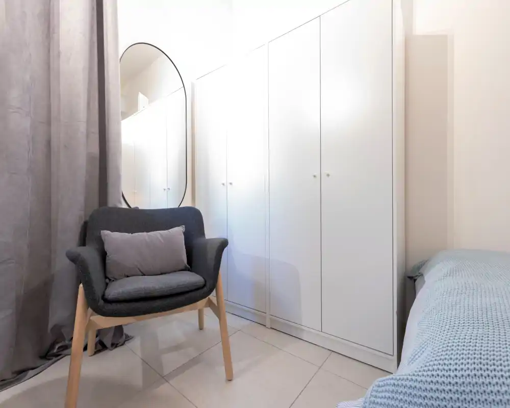 Rincón de dormitorio pequeño y luminoso con un sillón acogedor, armario y cama parcialmente visible con ropa de cama texturizada: estilo limpio y moderno, ideal para una sola persona.