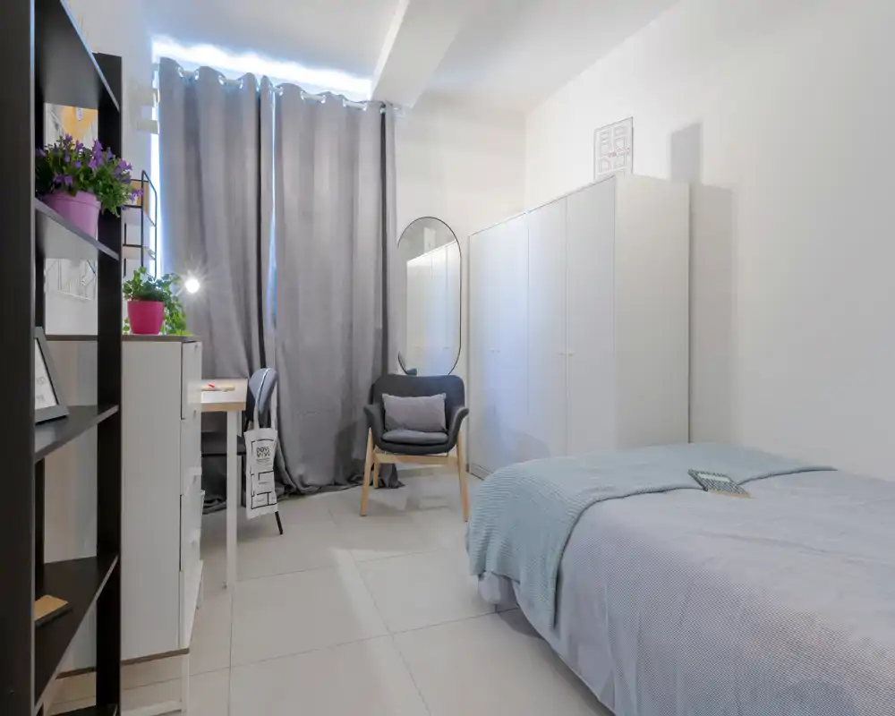 Dormitorio individual luminoso y ordenado con cama bien hecha, sillón confortable, armario y un pequeño escritorio junto a la ventana. Colores neutros y textiles suaves crean una atmósfera moderna y relajada.