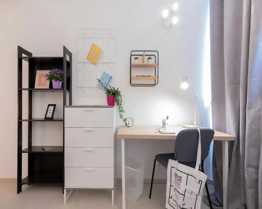 Rincón de dormitorio ordenado habilitado como pequeño espacio de trabajo: cómoda, estantería y escritorio con silla y lámpara. Tonos neutros, plantas y cortinas suaves generan un ambiente tranquilo.