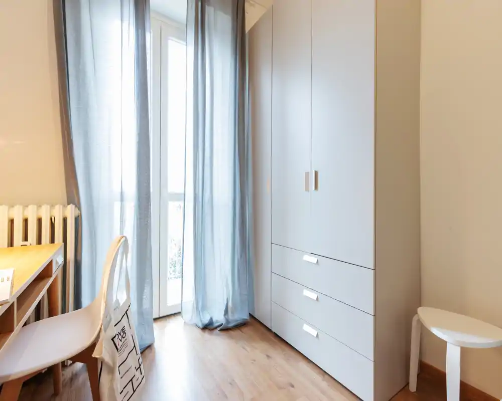 Bright bedroom corner with a large wardrobe, desk and chair by a balcony door dressed with light curtains; clean, minimal and softly lit.