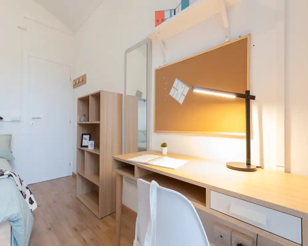 Bright, modern bedroom corner with a study desk, corkboard and bedside area. The compact bed and wooden shelving create a tidy, cozy student-style room ideal for work and rest.