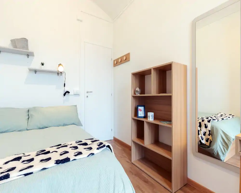 Bright, well-staged bedroom with a double bed dressed in soft green linens, a patterned throw, wall shelf with small decor, wooden shelving unit and a full-length mirror that adds depth.