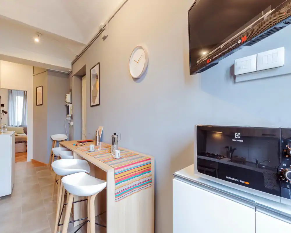 Compact modern kitchen area with a breakfast bar, three stools, countertop microwave and TV; bright, neatly styled space ideal for short-term stays.