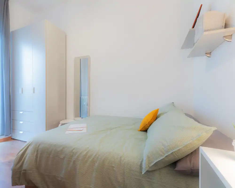 Chambre lumineuse et bien mise en scène avec un grand lit en linge vert pâle et un coussin jaune contrastant ; armoire et miroir en pied complètent l'espace chaleureux.