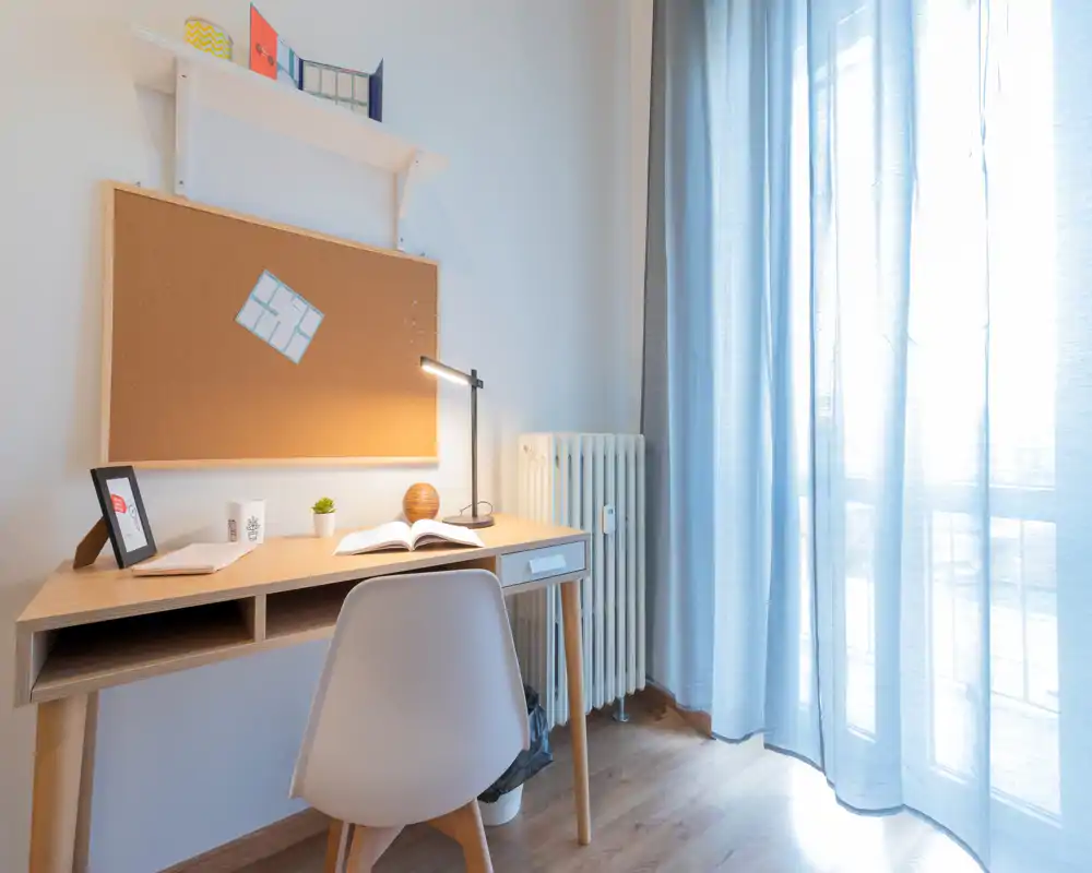 Coin bureau lumineux et minimaliste avec bureau en bois, chaise moderne, tableau en liège et lampe de bureau près d'une grande fenêtre aux rideaux légers.