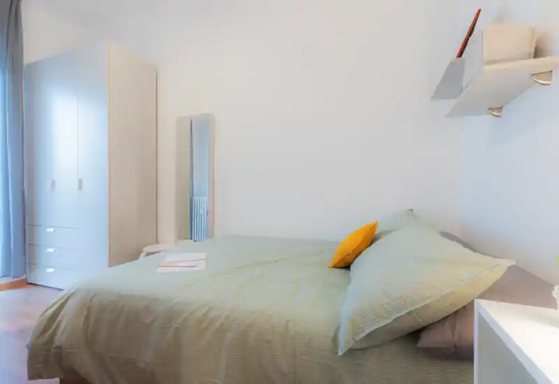 Chambre lumineuse et bien mise en scène avec un grand lit en linge vert pâle et un coussin jaune contrastant ; armoire et miroir en pied complètent l'espace chaleureux.