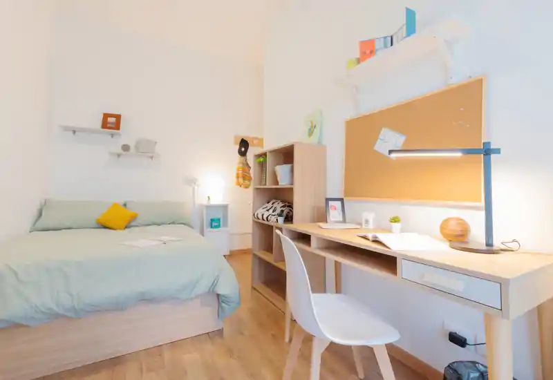 Chambre lumineuse et bien aménagée avec lit double, table de chevet et bureau fonctionnel avec panneau en liège — idéale pour étudiants ou professionnels recherchant une pièce cosy et pratique.