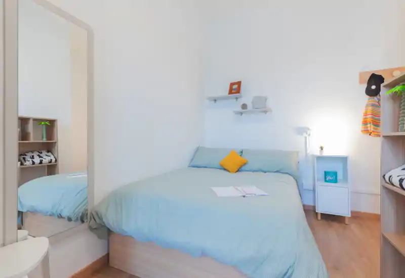 Chambre lumineuse et bien mise en scène avec un lit complet en linge bleu pâle, un coussin jaune contrastant, une table de chevet et des étagères décoratives — idéale pour une annonce locative.