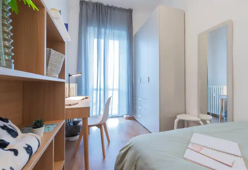 Chambre lumineuse et moderne avec lit bien fait, bureau près de la fenêtre et grande armoire — ambiance aérée avec une lumière naturelle douce filtrée par des voilages.