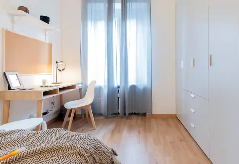 Chambre lumineuse et minimaliste avec un plaid à motif chaleureux, une armoire intégrée et un petit bureau en bois près de la fenêtre—parfaite pour un séjour calme et fonctionnel.