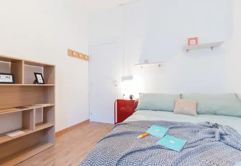 Chambre moderne et lumineuse avec un grand lit bien fait, coussins, plaid à motif et étagères en bois simples; parquet chaleureux et lampe de chevet minimaliste pour une ambiance accueillante.