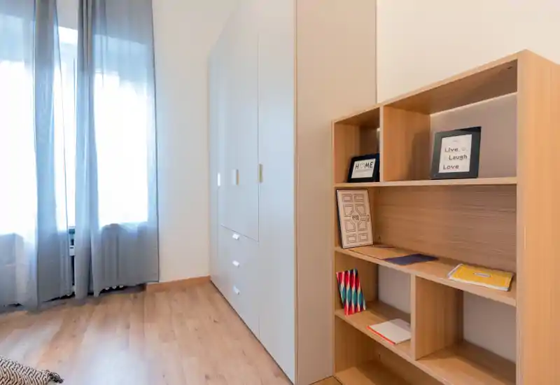 The image shows a storage area with a wardrobe and open shelving; no bed or clear living/kitchen elements are visible. The space appears tidy and well-lit with wooden floors and soft curtains.