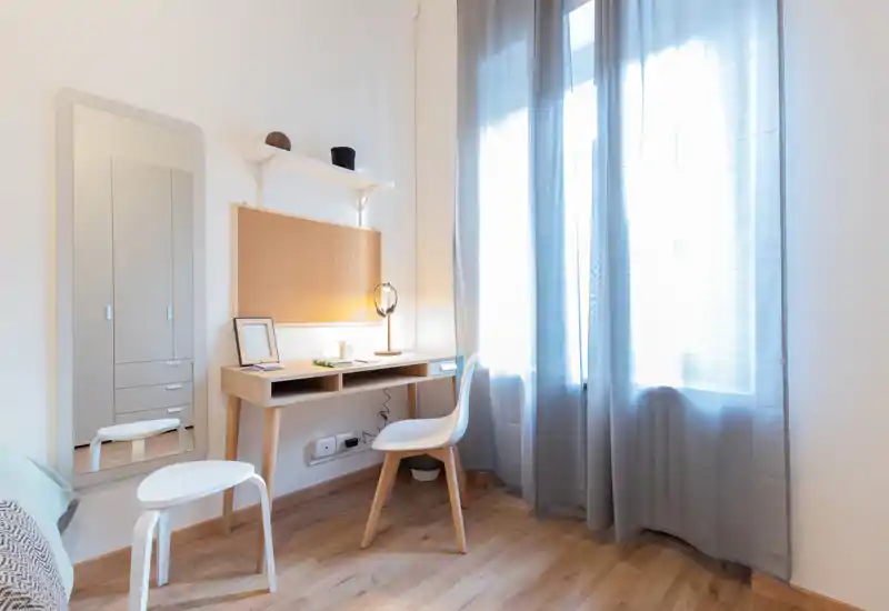 Small, bright bedroom corner with a study desk, chair and a partial view of the bed. Natural light from a large window, soft curtains and warm desk lighting create a comfortable workspace.