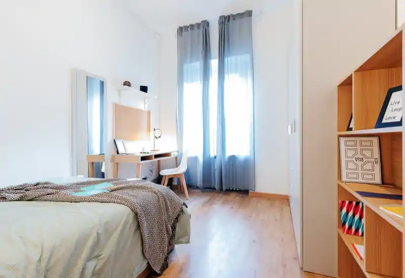 Chambre moderne et lumineuse avec lit bien fait, sol en bois, bureau près de la fenêtre et rideaux gris diffusant une belle lumière naturelle.