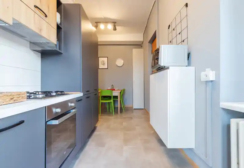 Modern galley kitchen with integrated oven, gas hob and a small dining nook with bright green chairs; clean lines and neutral tones create a fresh, functional space.