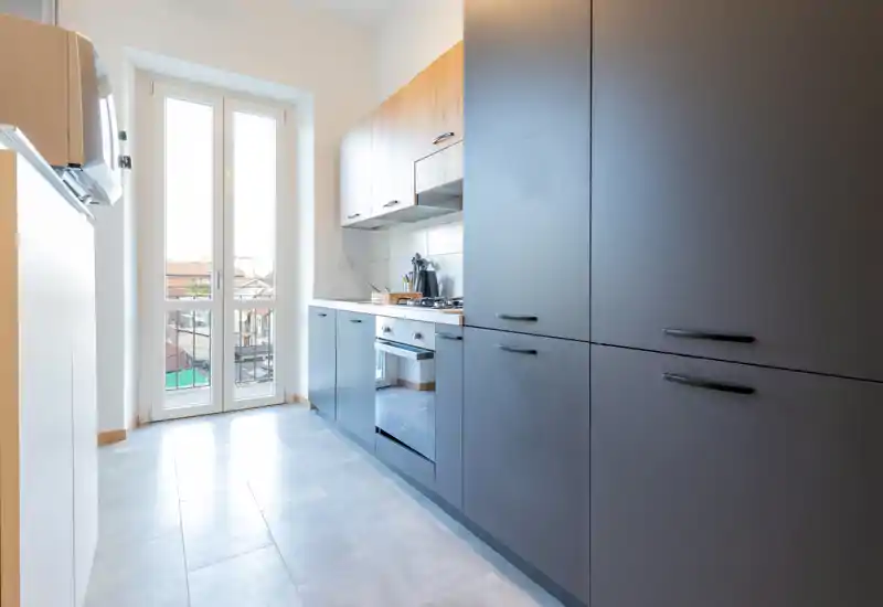 Modern galley kitchen with matte dark cabinets, wood upper cupboards, integrated oven and a tall window/door providing plenty of natural light.